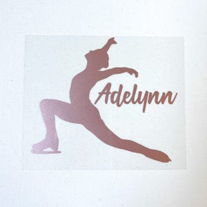 May include: A rose gold metallic decal featuring the silhouette of a figure skater in a lunge position with the name Adelynn written in a cursive script font. The design is displayed on a plain white textured surface.