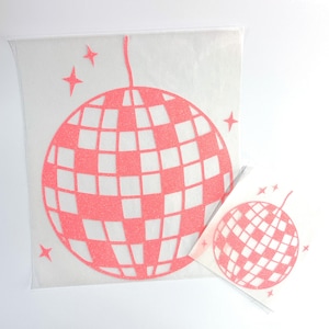 Puede incluir: Dos pegatinas de bola de discoteca rosas con una textura brillante. La pegatina más grande está en una funda de plástico transparente. Las bolas de discoteca tienen un patrón de cuadrícula y están rodeadas de estrellas.