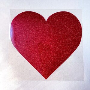 May include: A large, red glitter heart shape on a clear backing. The heart is a solid color with a sparkling texture. The background is a white canvas.