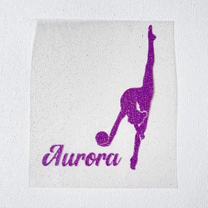 May include: A purple glitter iron-on transfer featuring a silhouette of a rhythmic gymnast performing a split with a ball. The name Aurora is written in a cursive script font below the gymnast, also in purple glitter on a white fabric background.
