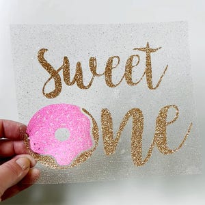May include: A glittery heat transfer vinyl design with the words "Sweet One" in gold script and a pink donut with gold sprinkles. The design is on a clear backing.