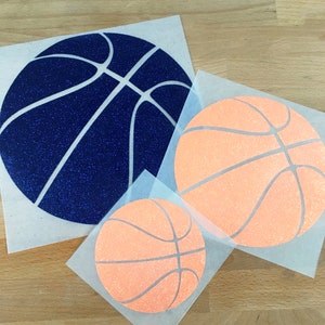 May include: Three basketball-themed decals in different sizes. One is navy blue with glitter, and the other two are orange with glitter. Each decal is on a square of transparent backing paper. Ideal for sports fans.