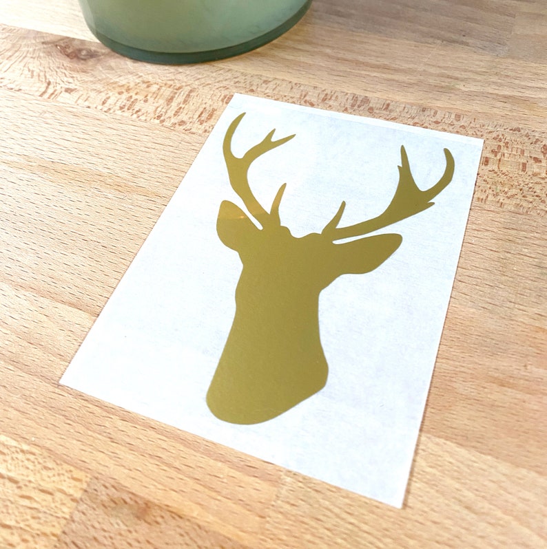 Op de afbeelding: Goudkleurig silhouet van een hertenkop op een witte achtergrond. De hertenkop heeft een groot gewei en is gecentreerd op de witte rechthoek. De sticker ligt op een houten oppervlak.