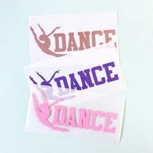 May include: Three decals with the word "DANCE" and a dancer silhouette. The top decal is a dusty rose color, the middle is purple, and the bottom is pink. The decals are on a white background with a light blue backdrop.