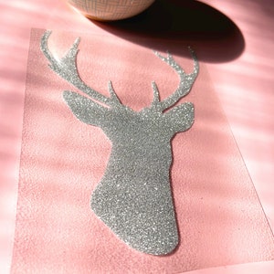 Op de afbeelding: Een zilveren glitter hertenkop ontwerp op een roze achtergrond. Een brandende kaars in een patroonkom staat op de achtergrond. De hertenkop is een uitgesneden vorm, waarschijnlijk voor knutselen of decoratie.