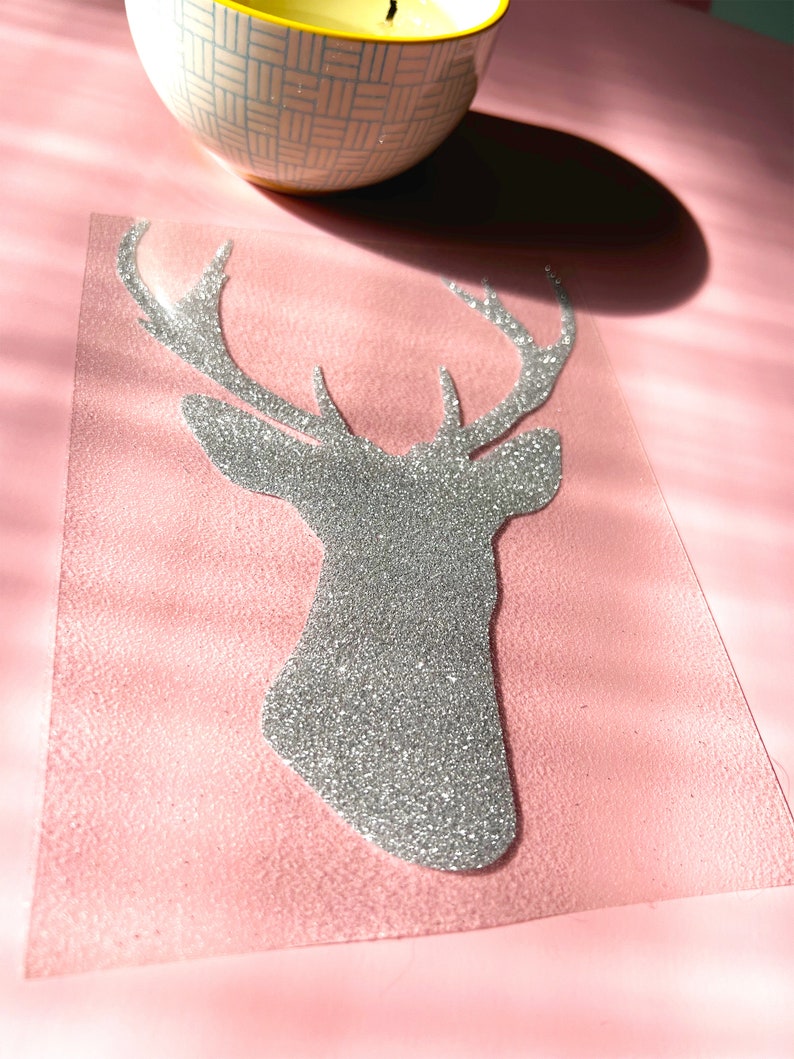 Op de afbeelding: Een zilveren glitter hertenkop ontwerp op een roze achtergrond. Een brandende kaars in een patroonkom staat op de achtergrond. De hertenkop is een uitgesneden vorm, waarschijnlijk voor knutselen of decoratie.