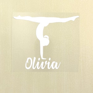 May include: A white vinyl decal featuring a silhouette of a gymnast performing a handstand with legs in a split position. Below the gymnast figure, the name Olivia is written in a white cursive script font on a clear adhesive backing.