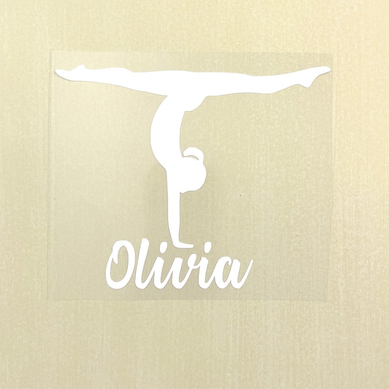 May include: A white vinyl decal featuring a silhouette of a gymnast performing a handstand with legs in a split position. Below the gymnast figure, the name Olivia is written in a white cursive script font on a clear adhesive backing.
