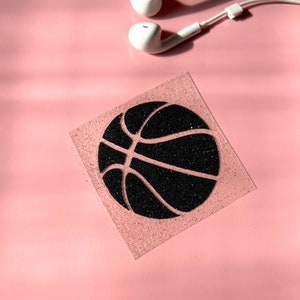 May include: A square, glittery pink coaster featuring a black basketball design. White earbuds are in the upper right corner, set against a solid pink background.