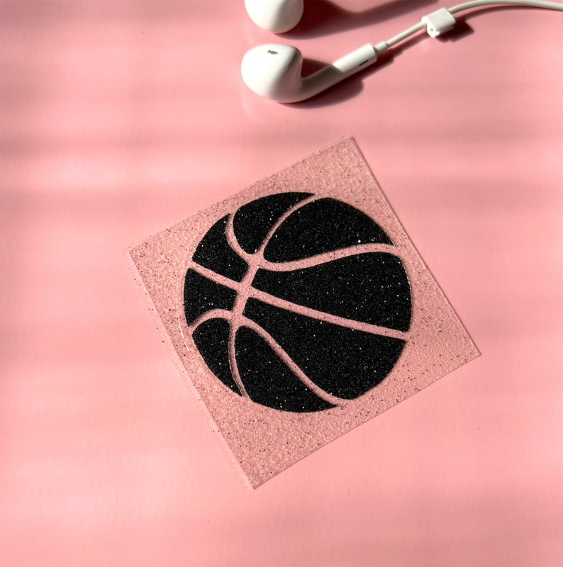 May include: A square, glittery pink coaster featuring a black basketball design. White earbuds are in the upper right corner, set against a solid pink background.