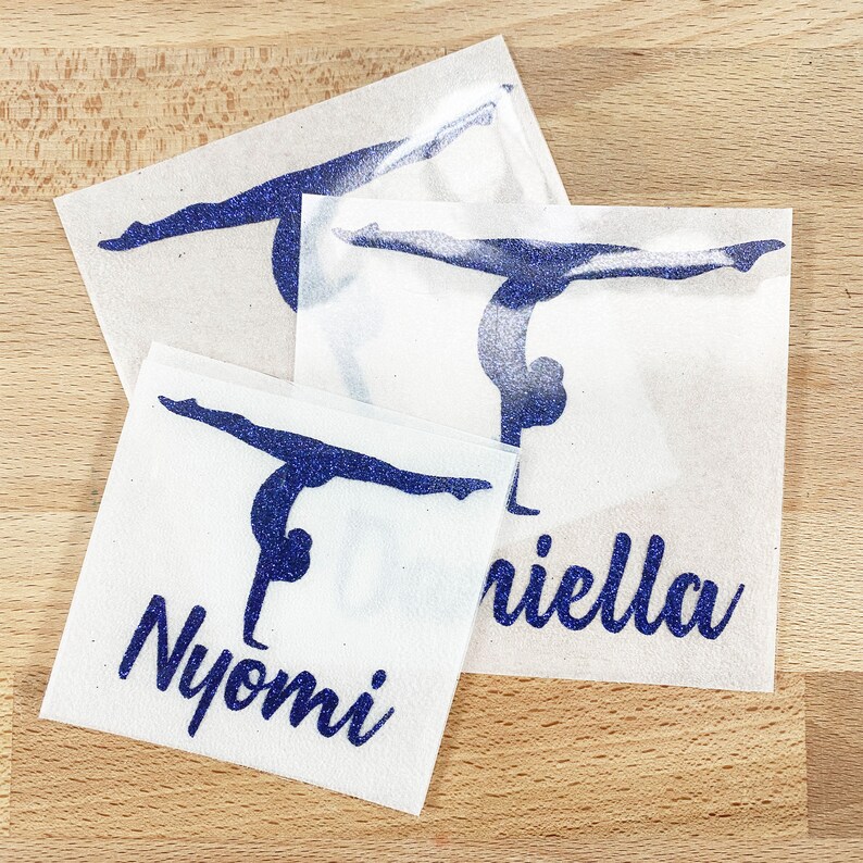 May include: Three square adhesive decals featuring a dark blue glitter gymnast silhouette in a handstand position. Two of the decals include the names Nyomi and Daniella printed in a dark blue glitter script font below the gymnast figure.