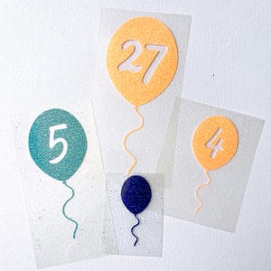 May include: Four balloon-shaped cutouts with glittery finishes on transparent sheets. One large orange balloon features the number 27, one teal balloon features the number 5, one small orange balloon features the number 4, and one small dark blue balloon.