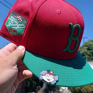 May include: A red and green baseball cap with a green "B" on the side and a patch that says "1981 World Champions Dodgers". The cap has a green brim and a cartoon character pin on the front.