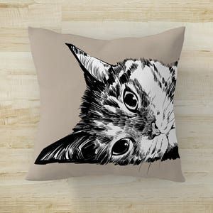 May include: A black and white illustration of a cat's face peeking out from behind a beige pillow. The cat's eyes are wide open and its whiskers are visible.