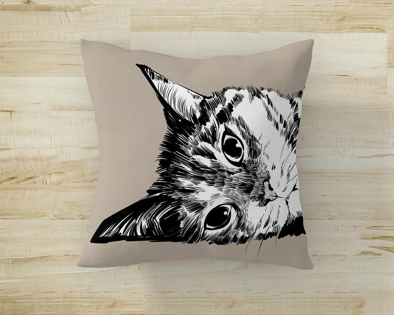 May include: A black and white illustration of a cat's face peeking out from behind a beige pillow. The cat's eyes are wide open and its whiskers are visible.