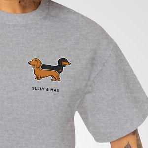May include: A light gray t-shirt featuring a cartoon illustration of two dachshunds, one brown and one black. The text "SULLY & MAX" is printed below the dogs. The shirt is made of a soft material.