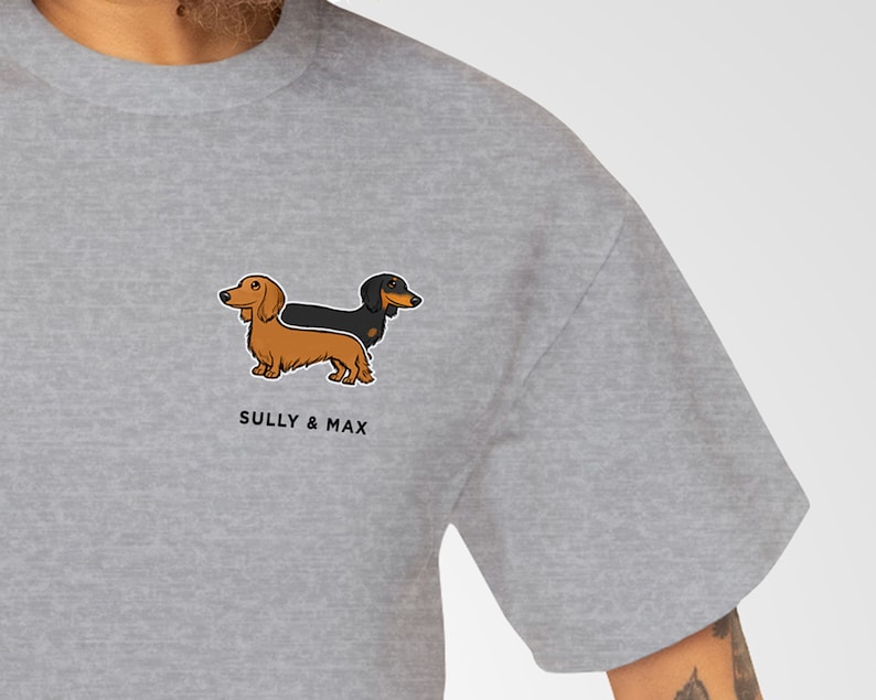 May include: A light gray t-shirt featuring a cartoon illustration of two dachshunds, one brown and one black. The text "SULLY & MAX" is printed below the dogs. The shirt is made of a soft material.