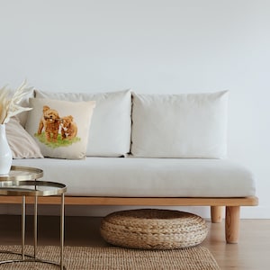 May include: A white couch with two beige pillows and a throw pillow featuring a watercolor illustration of two brown toy poodles playing with a stick. The couch is on a wooden frame with a woven wicker footstool in front of it.