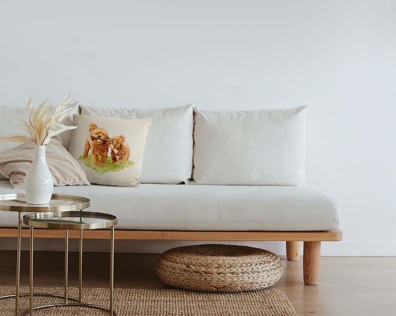 May include: A white couch with two beige pillows and a throw pillow featuring a watercolor illustration of two brown toy poodles playing with a stick. The couch is on a wooden frame with a woven wicker footstool in front of it.