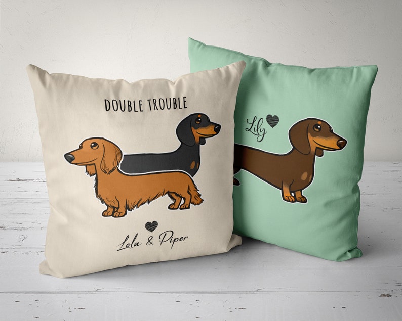 Puede incluir: Dos fundas de almohada de color crema con ilustraciones de perros salchicha. Una funda de almohada presenta un perro salchicha negro y marr&oacute;n con el texto "Double Trouble" y "Lola & Piper". La otra funda de almohada presenta un perro salchicha marr&oacute;n con el texto "Lily".