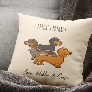 Puede incluir: Una almohada blanca con una ilustraci&oacute;n de dibujos animados de tres perros salchicha. El texto "Peter's Family" est&aacute; en la parte superior de la almohada. El texto "Leon, Hobbes, & Cooper" est&aacute; en la parte inferior de la almohada.
