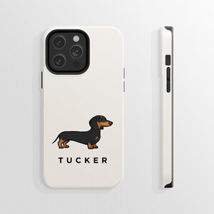 K&ouml;nnte beinhalten: Wei&szlig;e iPhone-H&uuml;lle mit einer Cartoon-Illustration eines schwarzen und braunen Dackels und dem Namen "TUCKER" darunter. Die H&uuml;lle hat eine schwarze Kamera-Erhebung mit drei Objektiven.