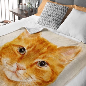K&ouml;nnte beinhalten: Eine weiche, pl&uuml;schige Kuscheldecke mit einer Aquarell-Illustration eines roten Katzenkopfes. Die Augen der Katze sind offen und schauen direkt auf den Betrachter. Die Decke ist hellbeige mit einer dezenten Textur.