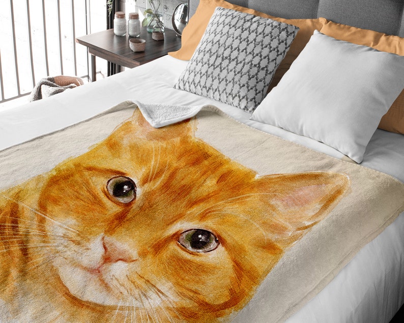 K&ouml;nnte beinhalten: Eine weiche, pl&uuml;schige Kuscheldecke mit einer Aquarell-Illustration eines roten Katzenkopfes. Die Augen der Katze sind offen und schauen direkt auf den Betrachter. Die Decke ist hellbeige mit einer dezenten Textur.