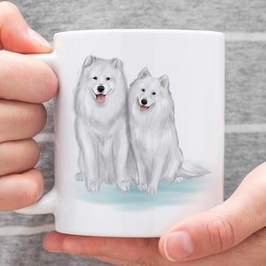K&ouml;nnte beinhalten: Wei&szlig;e Keramiktasse mit einer Aquarell-Illustration von zwei wei&szlig;en Samojeden, die nebeneinander sitzen.