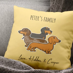 Puede incluir: Una almohada amarilla con el texto "Peter's Family" y una ilustraci&oacute;n de dibujos animados de tres perros salchicha. Los perros se llaman Leon, Hobbes y Cooper.