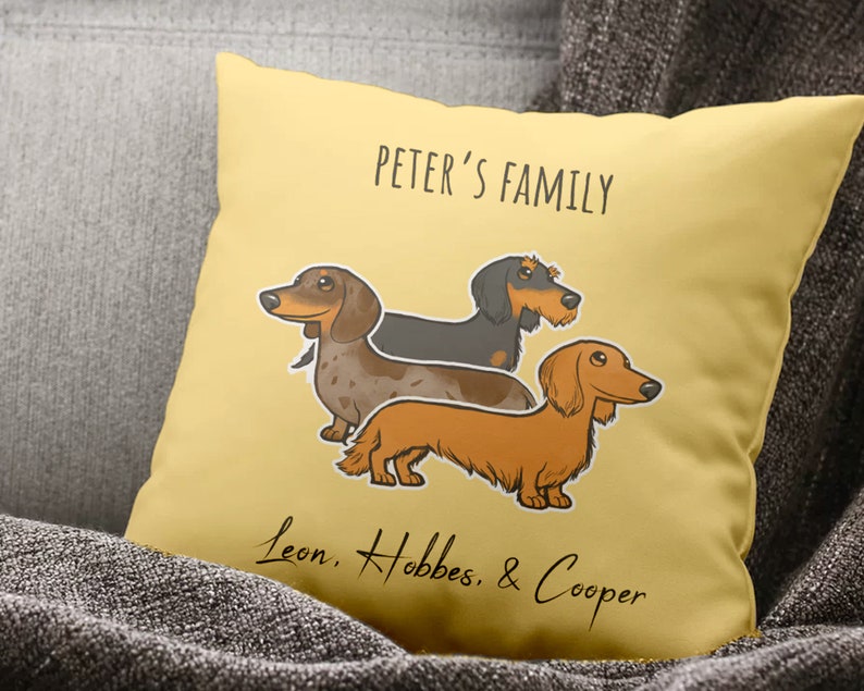 Puede incluir: Una almohada amarilla con el texto "Peter's Family" y una ilustraci&oacute;n de dibujos animados de tres perros salchicha. Los perros se llaman Leon, Hobbes y Cooper.