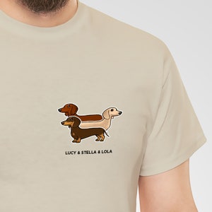 May include: Beige t-shirt featuring a cartoon illustration of three dachshund dogs in brown, tan, and cream colors. The text "LUCY & STELLA & LOLA" is printed below the dogs. The shirt is made of a soft, neutral-colored fabric.