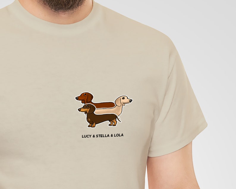 May include: Beige t-shirt featuring a cartoon illustration of three dachshund dogs in brown, tan, and cream colors. The text "LUCY & STELLA & LOLA" is printed below the dogs. The shirt is made of a soft, neutral-colored fabric.