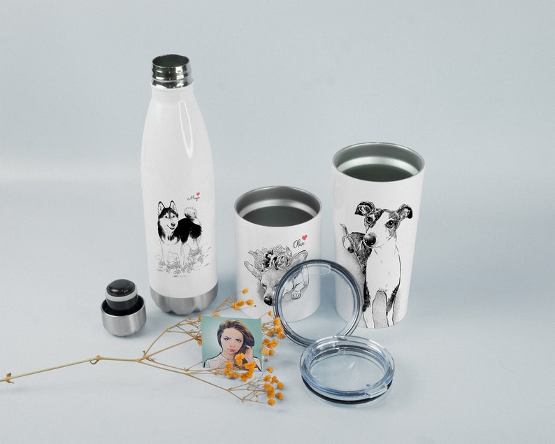 May include: Three white stainless steel tumblers with lids. The tumblers feature black and white illustrations of dogs and a deer. One tumbler has the name "Oliver" on it. The tumblers are on a white surface with a small bouquet of yellow flowers.