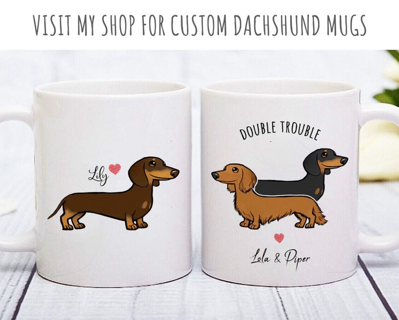 Puede incluir: Dos tazas de cer&aacute;mica blanca con ilustraciones de dachshund. Una taza presenta un dachshund marr&oacute;n con el nombre "Lily" y un coraz&oacute;n. La otra taza presenta un dachshund negro y marr&oacute;n con el texto "Double Trouble" y los nombres "Lala & Piper" con un coraz&oacute;n.