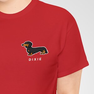 May include: A red t-shirt featuring a cartoon illustration of a black and brown dachshund dog. The dog is positioned on the lower right side of the shirt, with the name "DIXIE" printed in white below it. The shirt has short sleeves.
