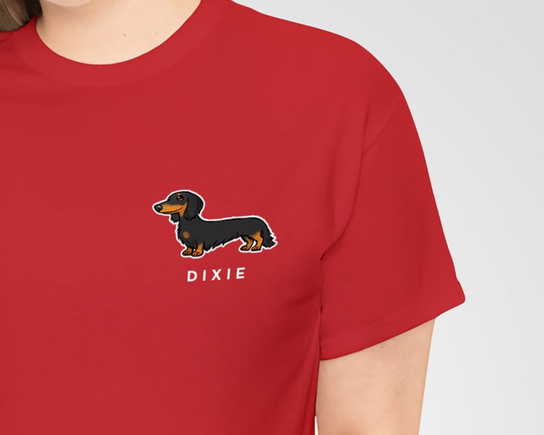 May include: A red t-shirt featuring a cartoon illustration of a black and brown dachshund dog. The dog is positioned on the lower right side of the shirt, with the name "DIXIE" printed in white below it. The shirt has short sleeves.