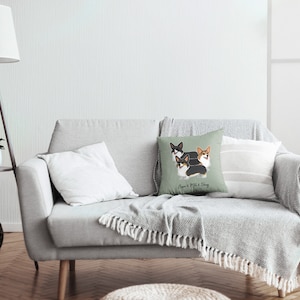 May include: A light gray couch with a throw blanket and three throw pillows. The center pillow features a green background with a cartoon illustration of three corgis and the text "Corgi & Whisk & Daisy".