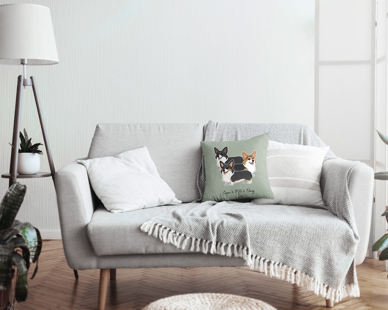 May include: A light gray couch with a throw blanket and three throw pillows. The center pillow features a green background with a cartoon illustration of three corgis and the text "Corgi & Whisk & Daisy".