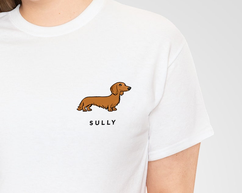 May include: White t-shirt featuring a cartoon illustration of a brown dachshund dog. The dog is standing, with the name "SULLY" printed in black below it. The shirt is made of a soft, comfortable material.