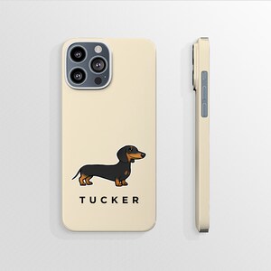 K&ouml;nnte beinhalten: Eine cremefarbene iPhone-H&uuml;lle mit einer Illustration eines schwarzen und braunen Dackels und dem Namen "TUCKER" unten aufgedruckt. Die H&uuml;lle hat einen Kameraausschnitt und einen seitlichen Knopf.