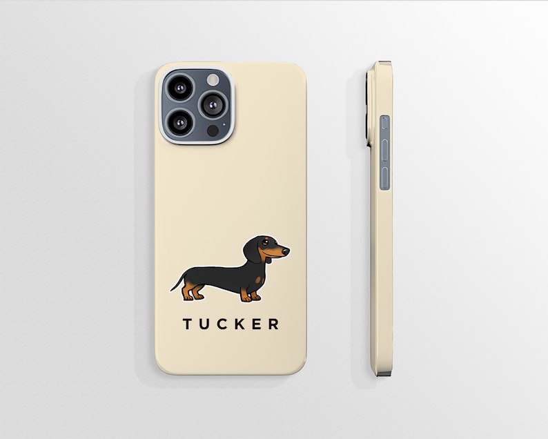 K&ouml;nnte beinhalten: Eine cremefarbene iPhone-H&uuml;lle mit einer Illustration eines schwarzen und braunen Dackels und dem Namen "TUCKER" unten aufgedruckt. Die H&uuml;lle hat einen Kameraausschnitt und einen seitlichen Knopf.