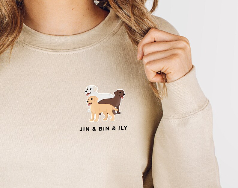 Pode incluir: Uma camisa de moletom bege com um gr&aacute;fico de tr&ecirc;s labradores retrievers em diferentes cores. O texto "JIN & BIN & ILY" &eacute; impresso abaixo do gr&aacute;fico.