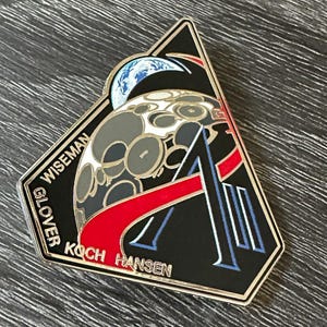 May include: A triangular black and silver enamel pin featuring a stylized moon with a blue and white Earth in the background. The pin has the names WISEMAN, GLOVER, KOCH, and HANSEN around the edges.