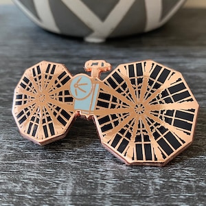 May include: A rose gold enamel pin featuring a stylized depiction of a satellite with two large, black, octagonal antennas. The antennas have a radial pattern of lines.