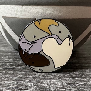 Op de afbeelding: Een ronde emaille pin met een kleurrijk abstract ontwerp. De pin heeft een bruin, paars, geel en wit ontwerp.