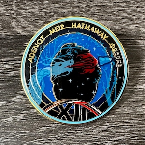 May include: A round, blue, and gold enamel pin with the text "ADENOT MEIR HATHAWAY DEGEE" around the edge. The pin features a stylized image of a dragon and a space scene. The number "XII" is at the bottom.