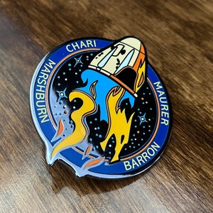 May include: A blue and black enamel pin with a rocket ship launching into space. The names "Chari", "Marshburn", "Barron", and "Maurer" are written around the edge of the pin.
