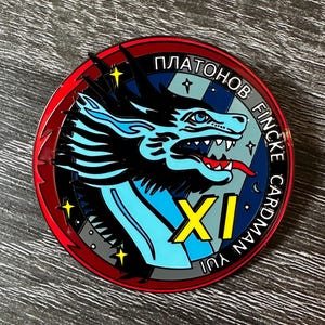 May include: A round, colorful metal pin featuring a stylized blue dragon with a red tongue and sharp teeth. The pin has text in multiple languages, including "PLATONOV FINCKE CARDMAN YUI XI". The border is red and black.