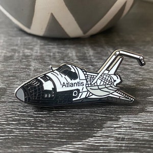 以下が含まれることがあります： スペースシャトルアトランティスの白黒イラストが描かれた銀色のエナメルピン。ピンには「アトランティス」という文字が側面に印刷されています。